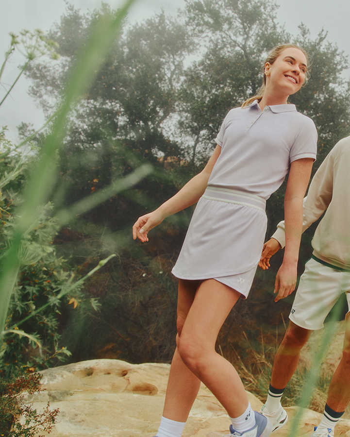 A man and a woman walking hand-in-hand outdoors in a natural, grassy landscape. The woman is wearing a light lavender-gray short-sleeve polo shirt and matching shorts, while the man wears a light beige hoodie, matching shorts, and white sneakers.
